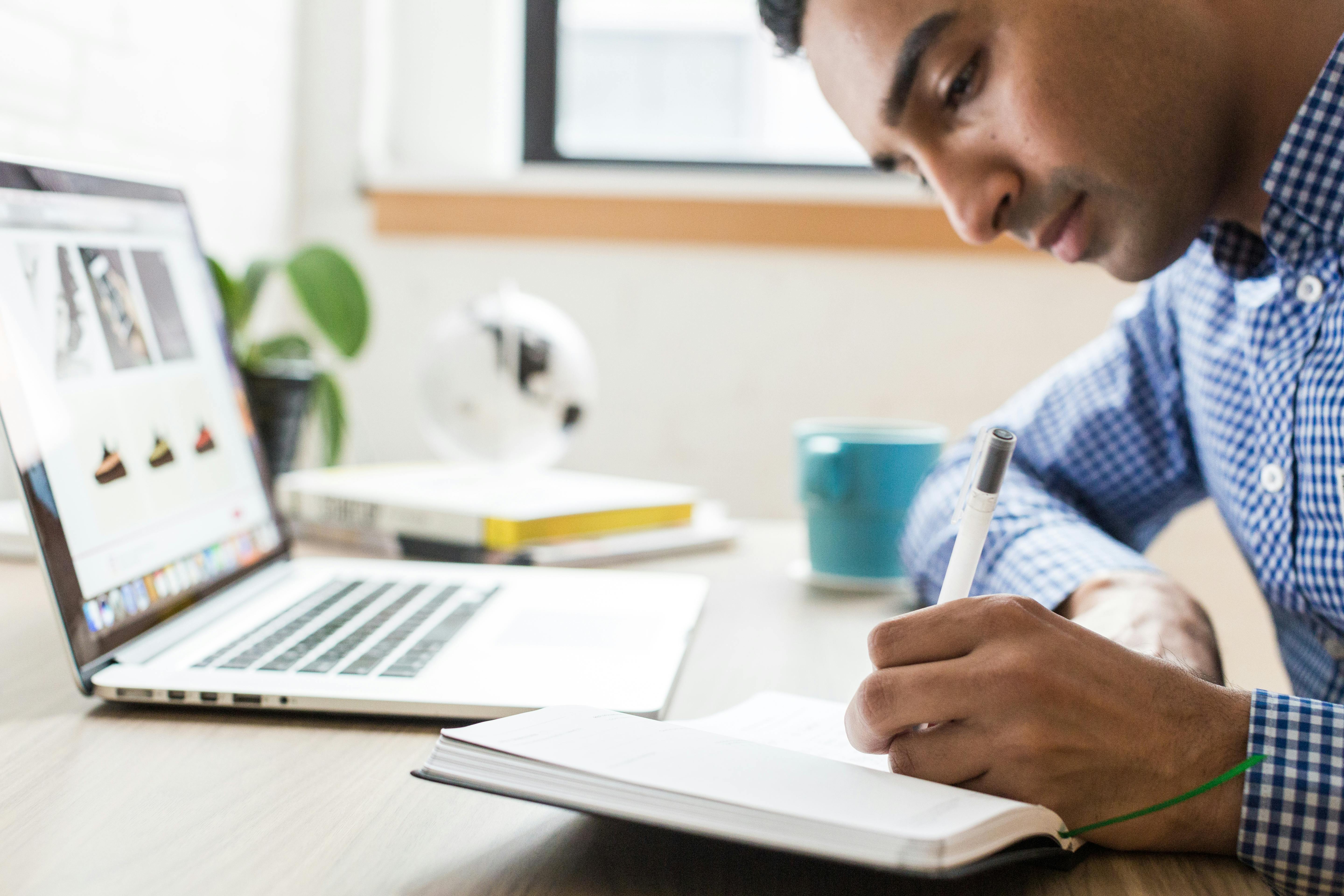 Man behind a laptop writing notes in his notebook