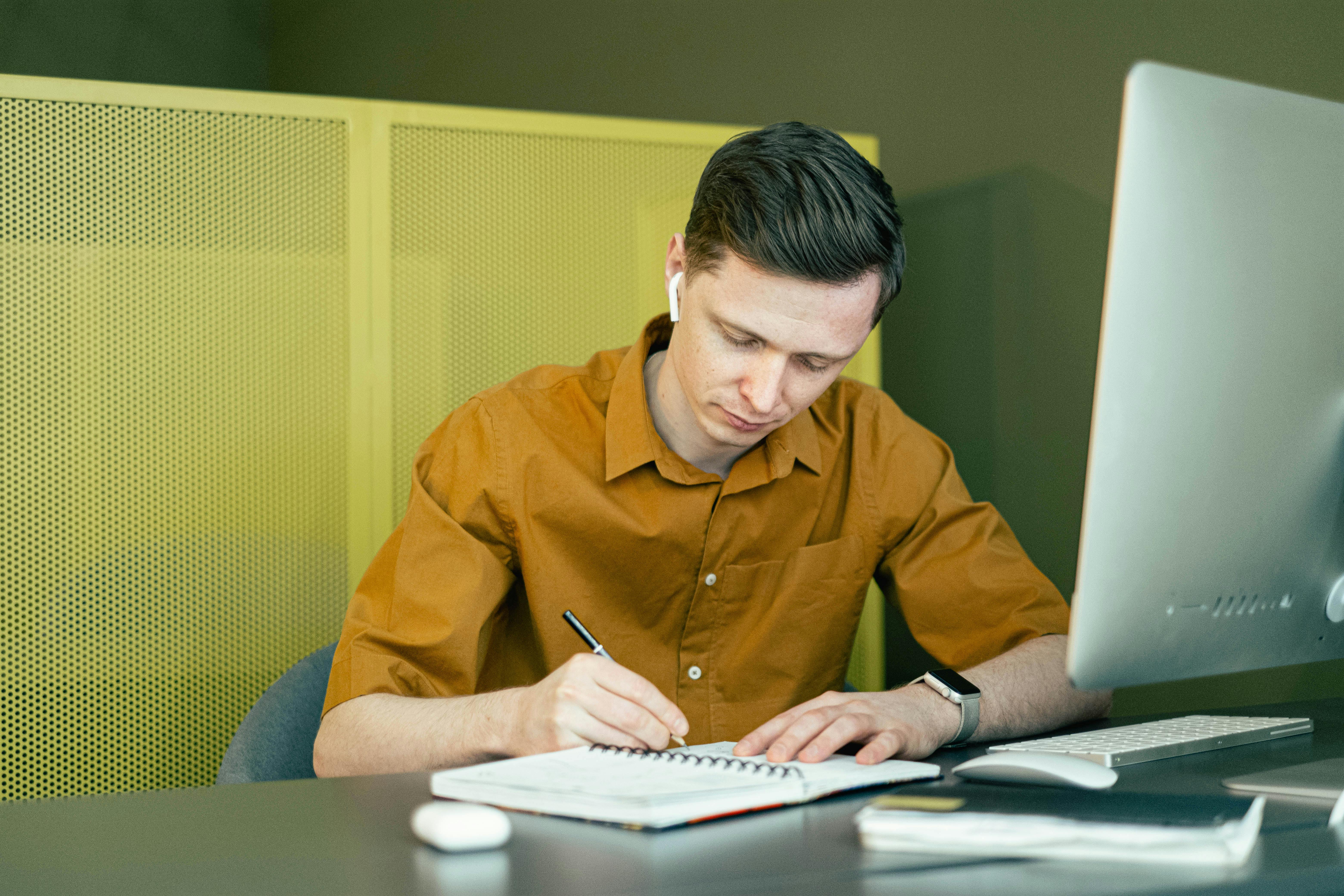 Young man sat behind his computer taking notes in a notebook