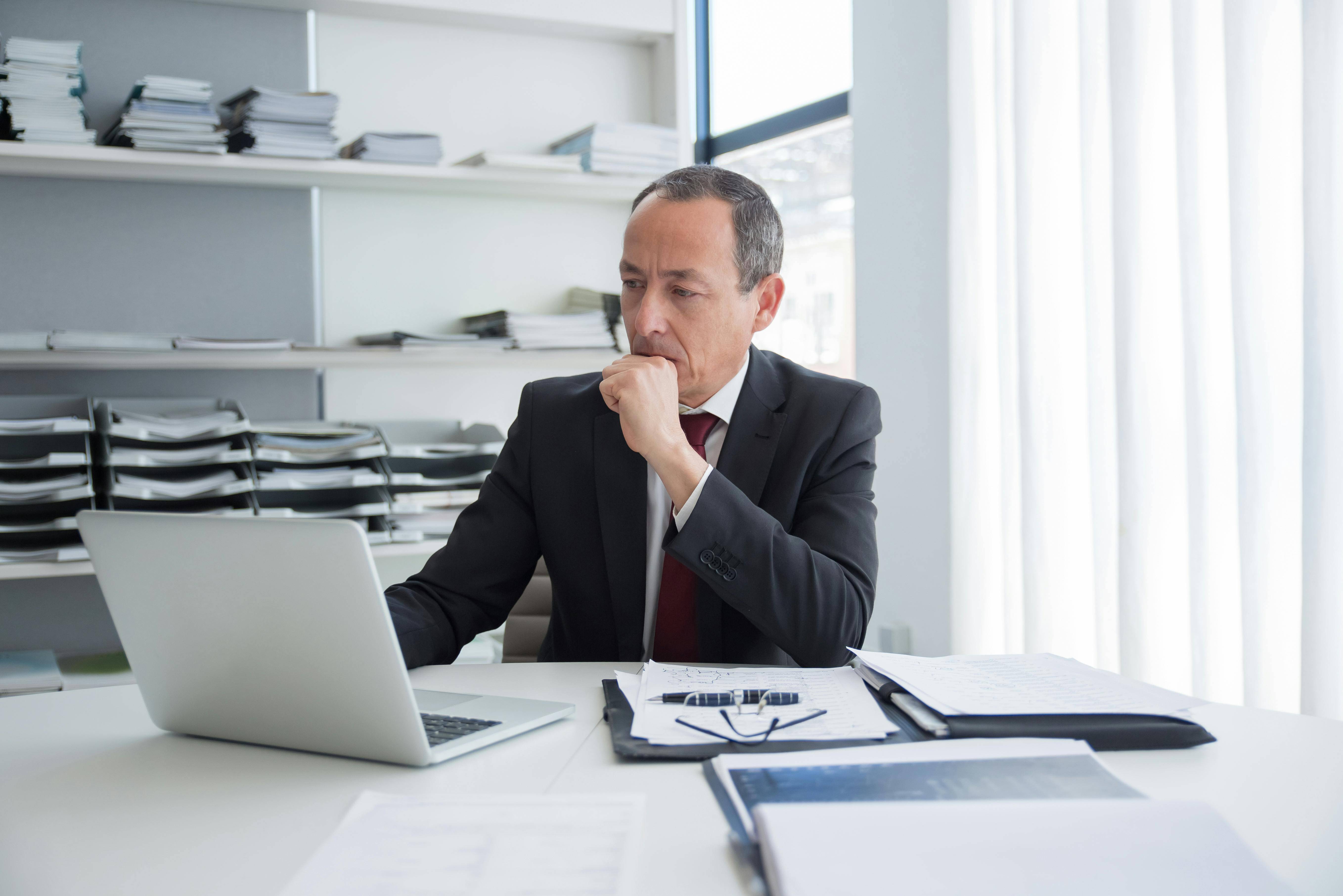 Man at his desk surrounded by documents looking at his laptop